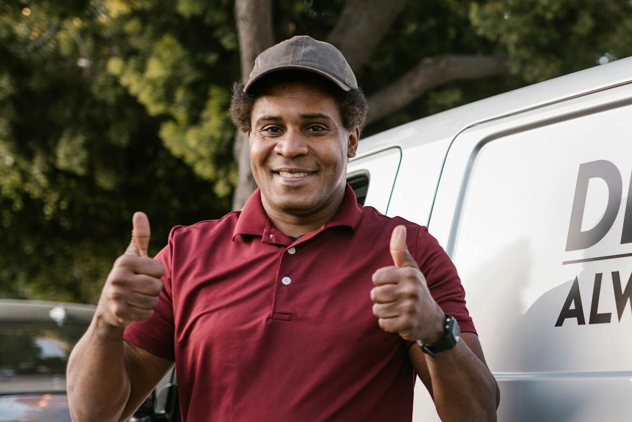 Happy deliveryman in maroon shirt giving thumbs up by delivery van outdoors.